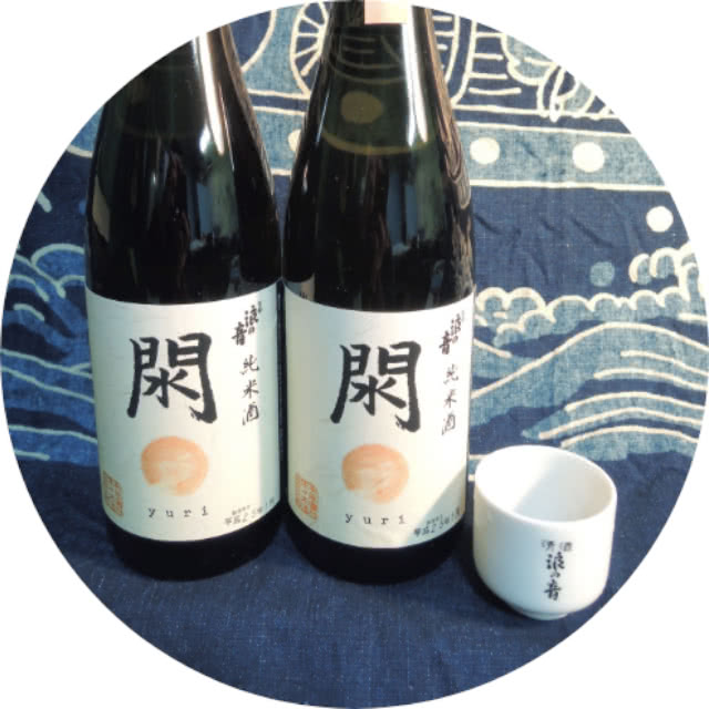 酒「閖」の瓶が二本と、日本酒用のぐい呑み。和風の布が背景に敷かれている。