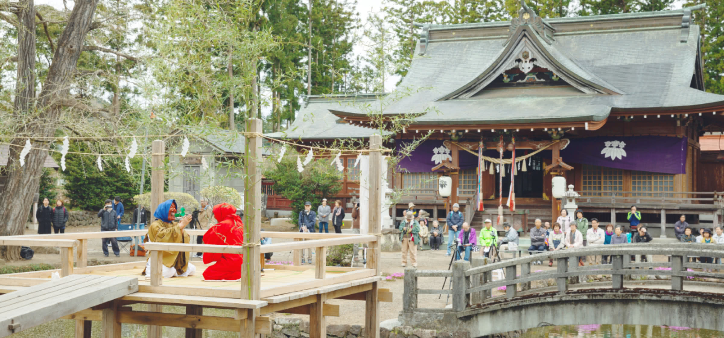 日本の伝統的な神社と木製の橋、舞台にはカラフルな衣装を着た二人の演者が座り、観客が見守っている。